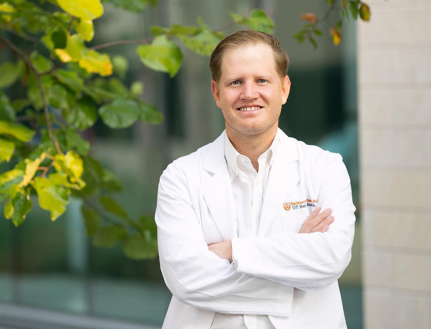 Endocrine surgeon Samuel E. "Rhett" Long, MD, FACS, posing next to a tree outside of the Health Discovery Building in downtown Austin. His arms are crossed and he is wearing a white coat with an embroidered UT Health Austin logo.