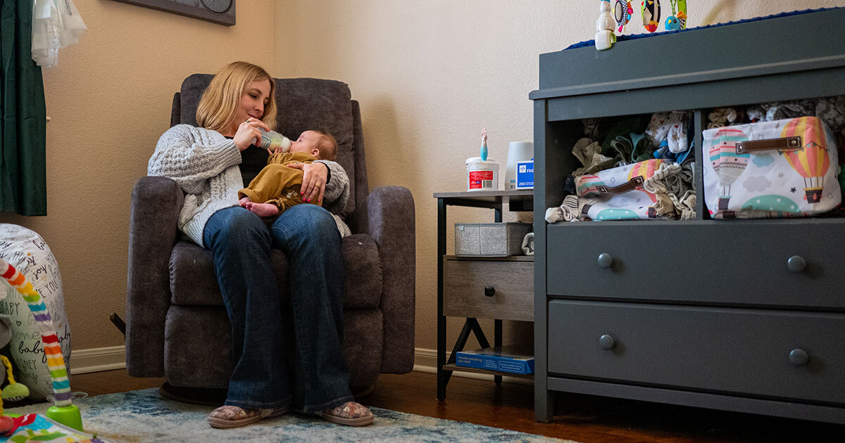 Woman sitting in a nursery feeding a baby with a bottle while seated in a cushioned recliner next to a dresser filled with baby supplies.