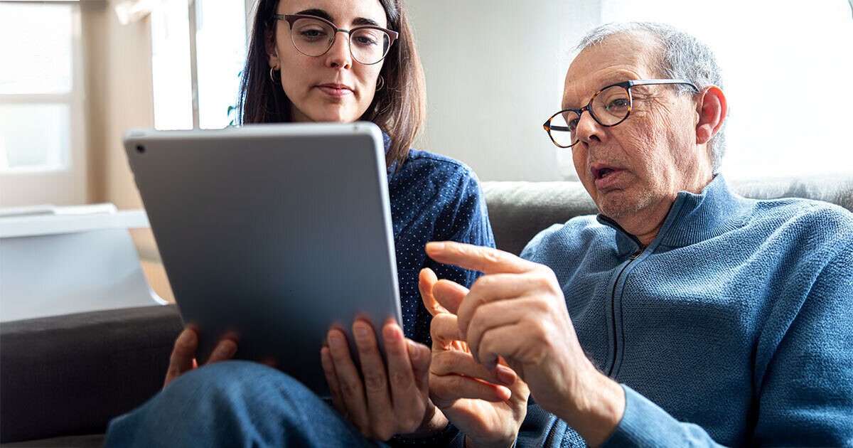 Older man and younger woman sitting on a couch, looking at a tablet together, symbolizing the use of technology to support cognitive health in older adults.