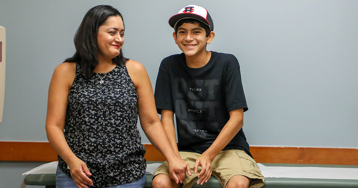 Joaquin, a pediatric cardiology patient who underwent open heart surgery to correct a congenital defect, sits on a table next to his mother, holding her hand.