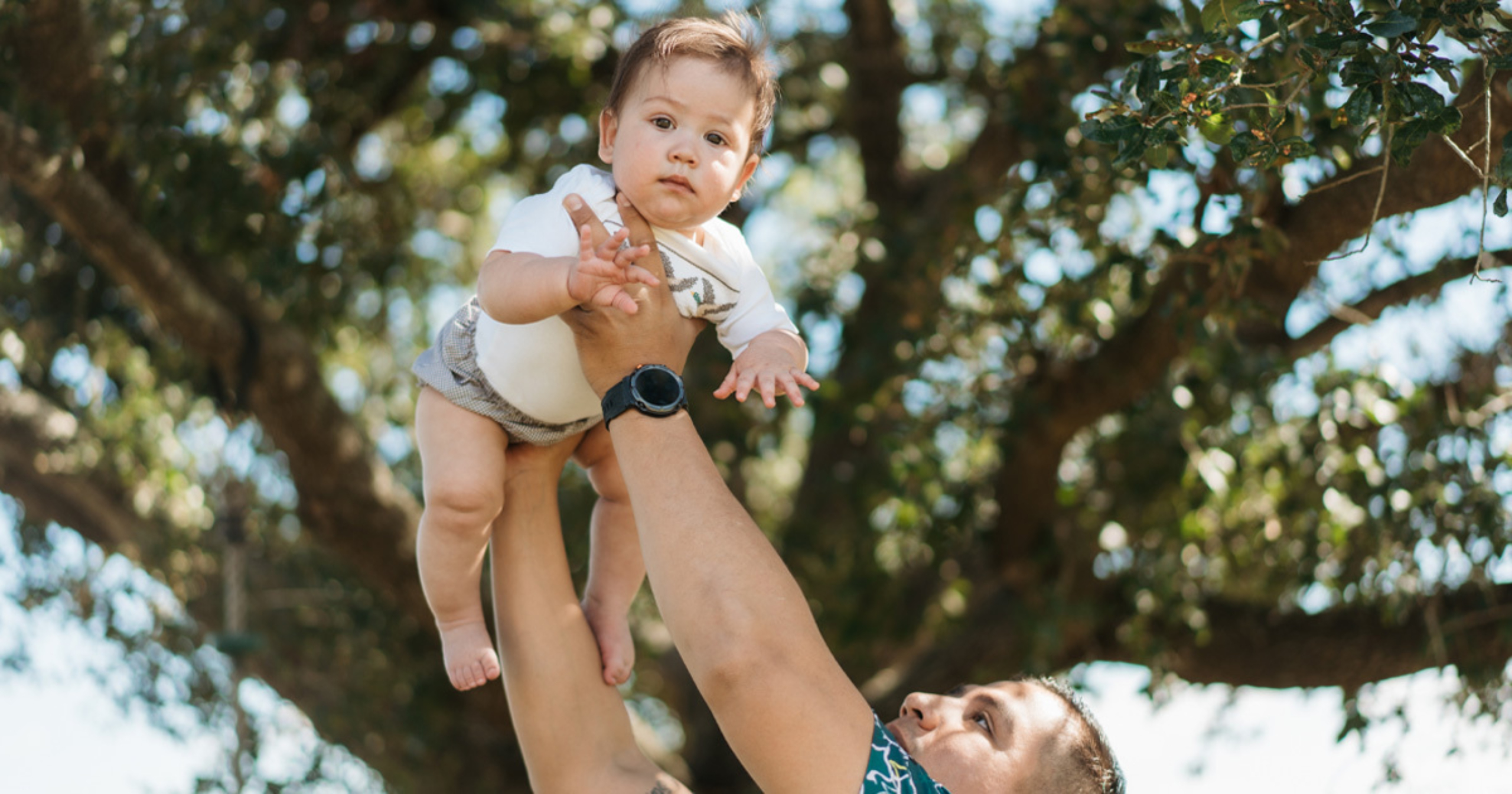 Man lifting a baby into the air outdoors under a large tree, with the baby looking directly at the camera.