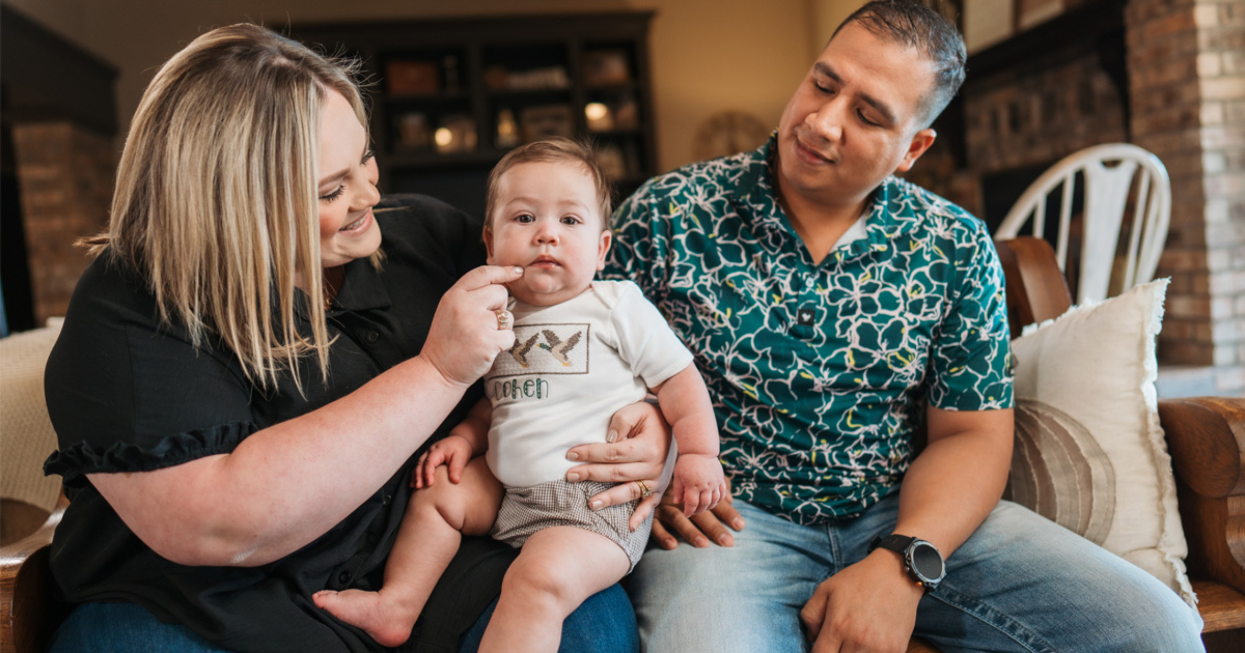 Smiling parents sitting on a couch with their baby, who wears a personalized shirt with the name "Cohen," as the mother gently touches the baby’s cheek.