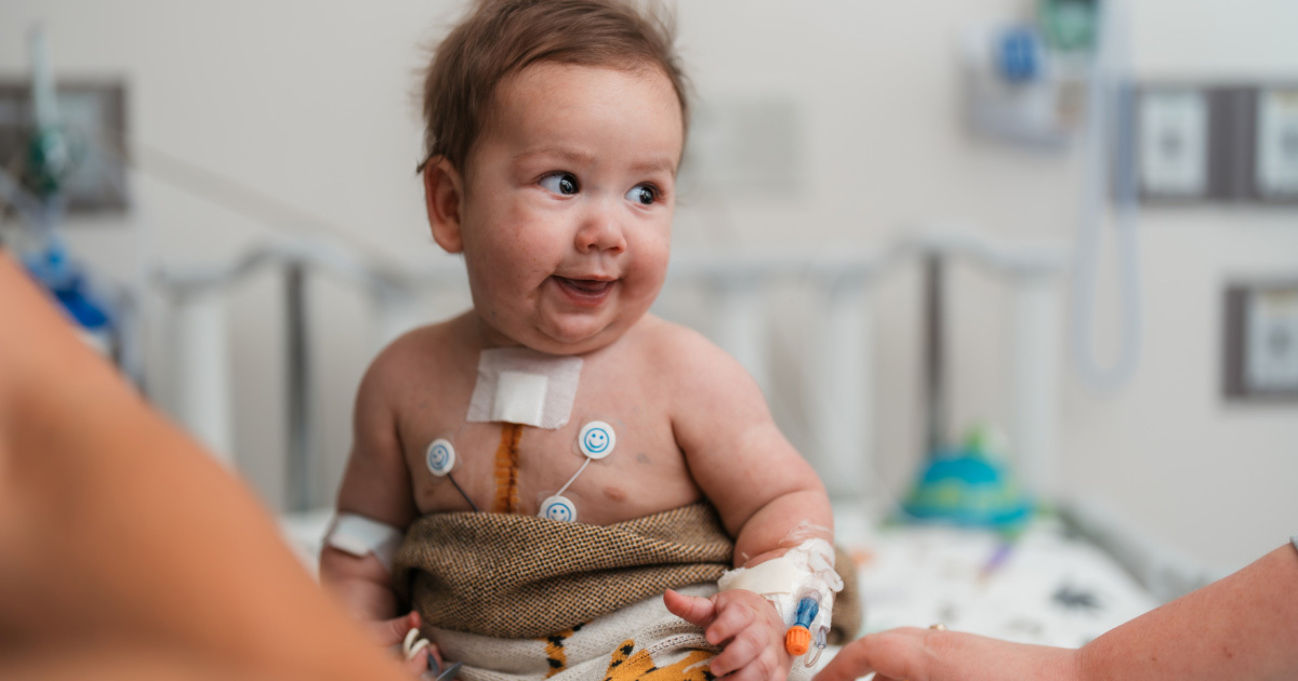 Smiling baby sitting up in a hospital crib with medical monitors attached to their chest and an IV in their arm, wrapped in a blanket after surgery.