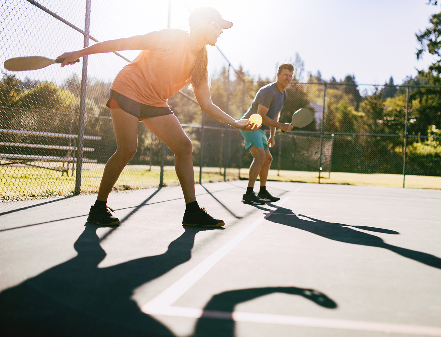 Two people playing pickleball on an outdoor court, with one player lunging forward to hit the ball, promoting tips for preventing common pickleball injuries.