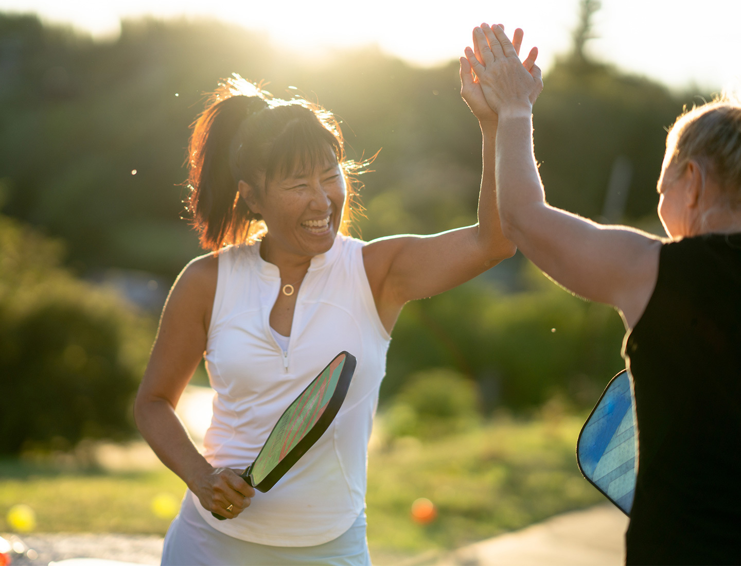 Two adult women in athletic wear high-five while holding pickleball paddles outdoors in the late afternoon sun.