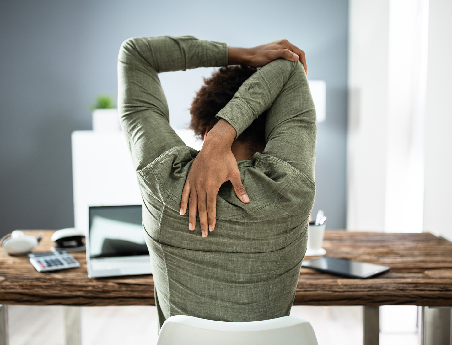 The backside of a woman wearing a forest green shirt is shown as she's sitting at a work desk and stretching. Her left arm reaches over her head and grabs her right elbow to stretch her triceps.
