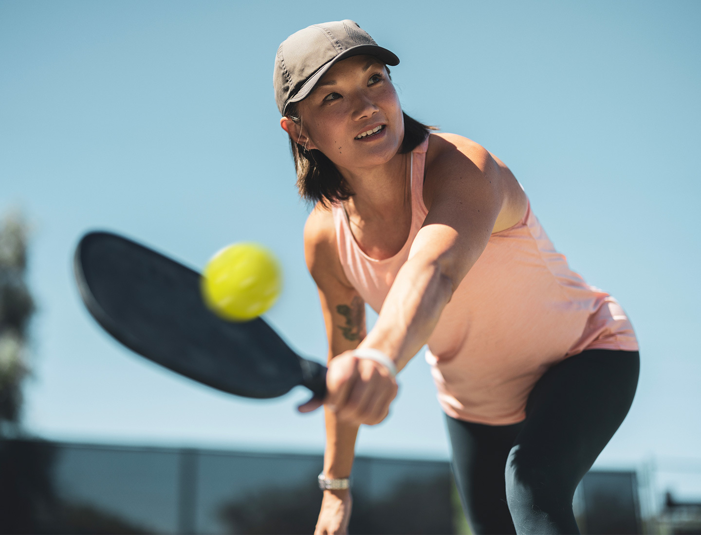 Adult woman in athletic wear and cap swings a paddle to hit a pickleball outdoors on a sunny day.