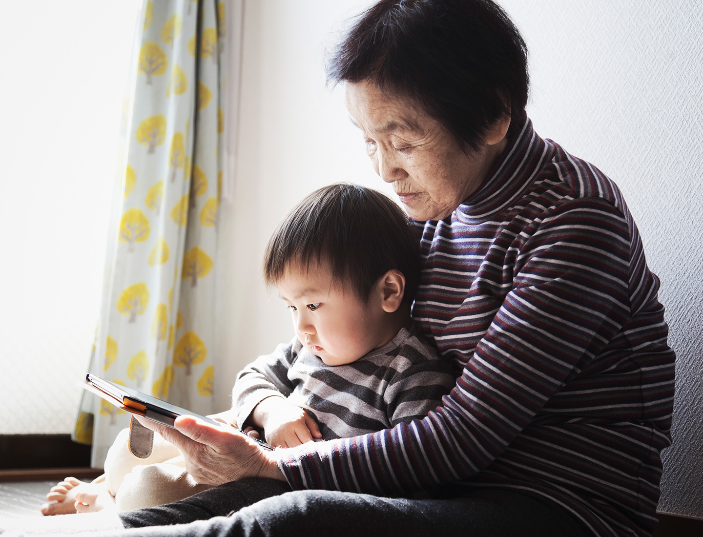 An Asian grandmother holds her grandson in her lap. She holds a mobile phone at which both of them are looking.