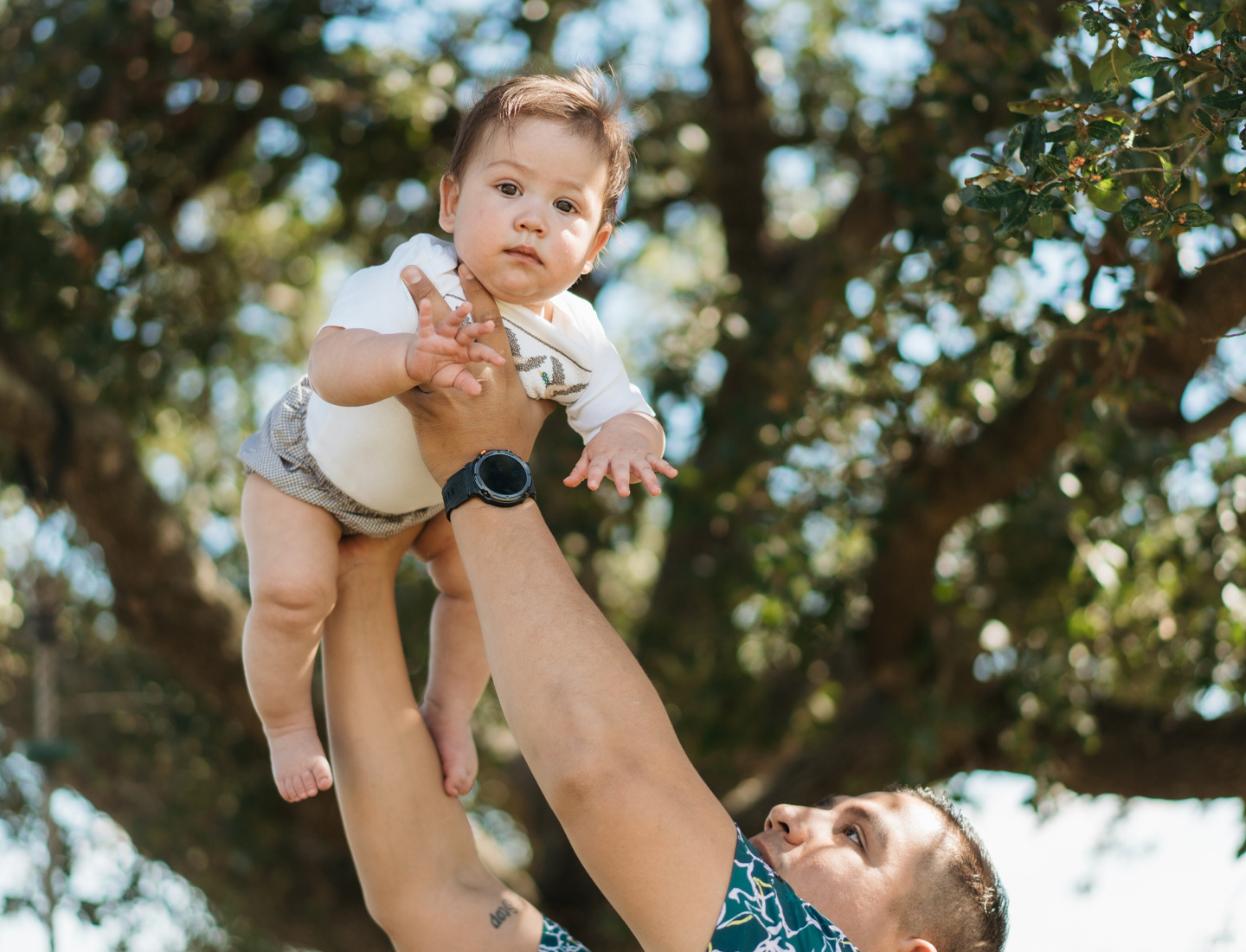 Man lifting a baby into the air outdoors under a large tree, with the baby looking directly at the camera.
