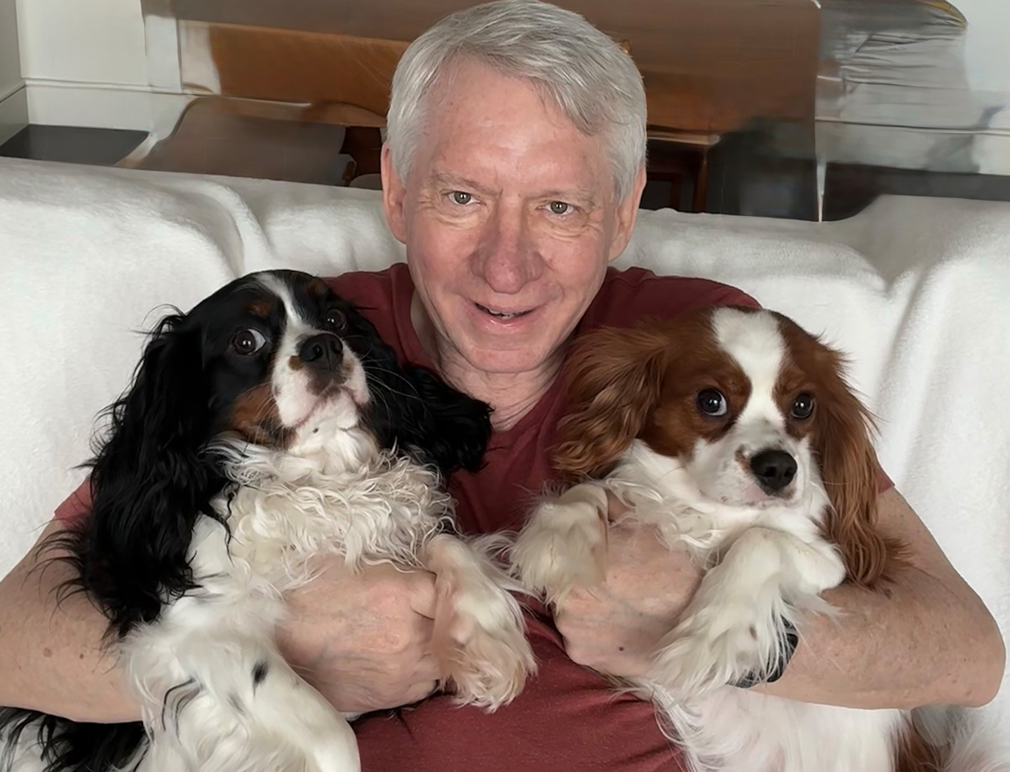 Smiling Donald “Don” Kettl, with short white hair sitting on a couch, holding two Cavalier King Charles Spaniels — one with black and white fur and one with chestnut and white fur.