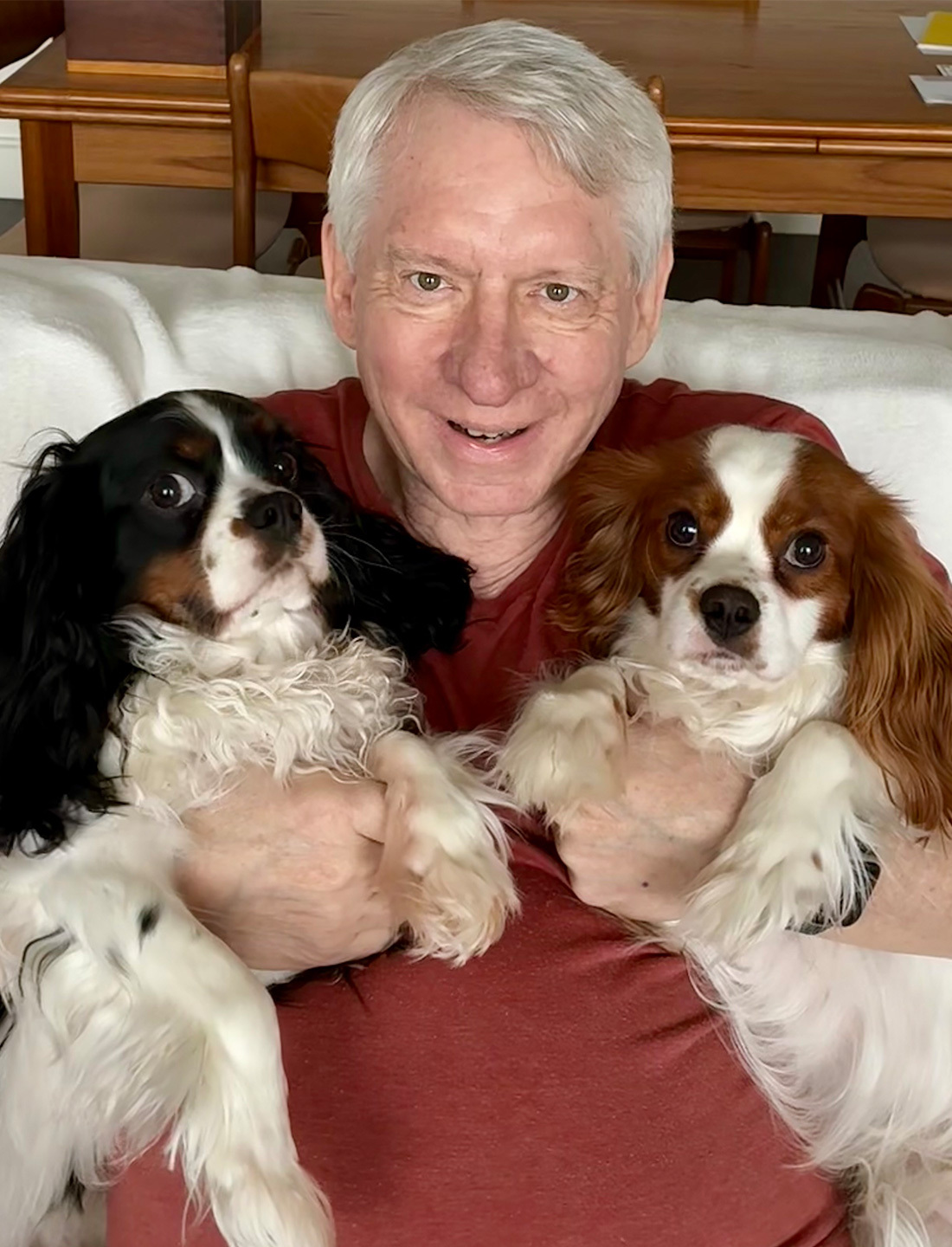 Smiling Donald “Don” Kettl, with short white hair sitting on a couch, holding two Cavalier King Charles Spaniels — one with black and white fur and one with chestnut and white fur.