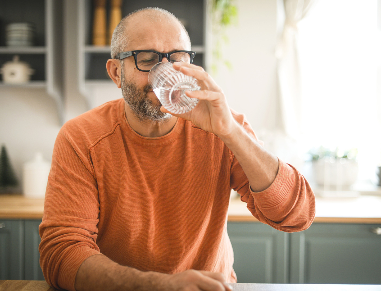 A man in an orange shirt drinks a glass of water in his kitchen.
