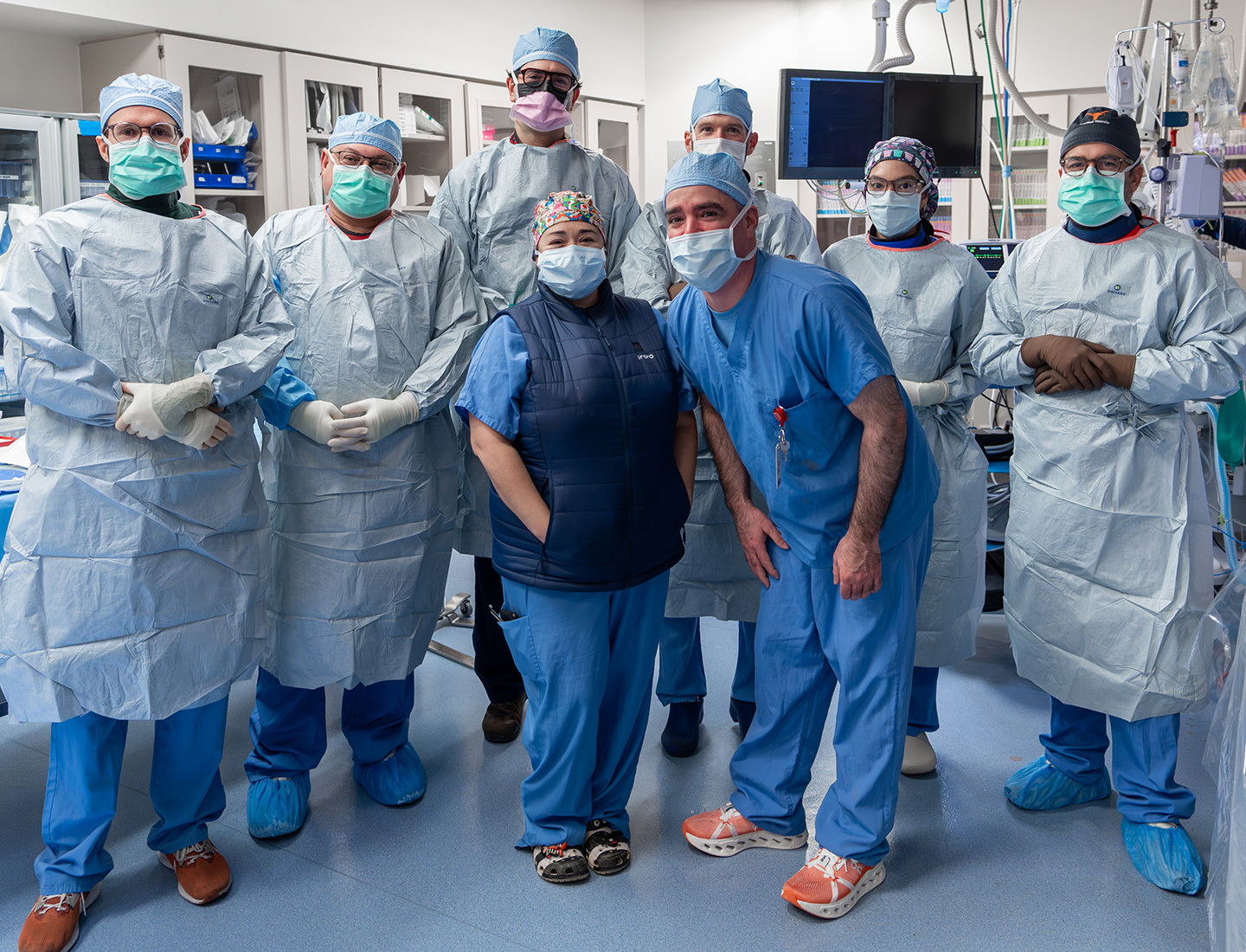 Cardiovascular care team in scrubs and masks pose together in a procedure room.