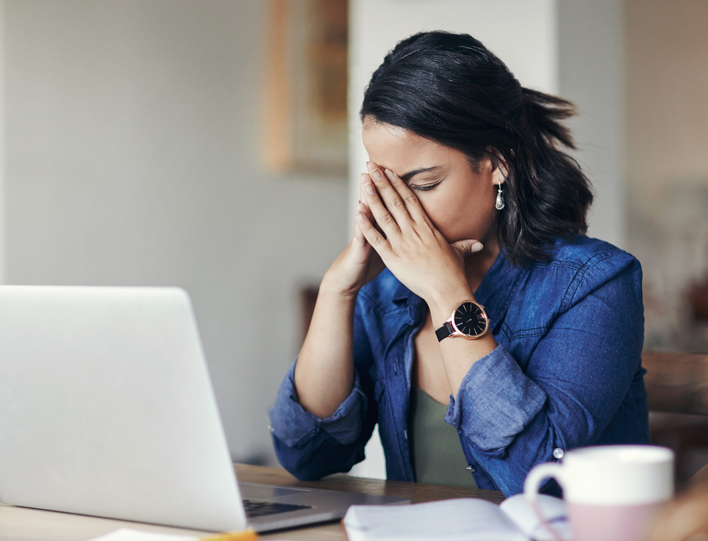 A woman sitting at a table with a laptop cradles her face in her hands.