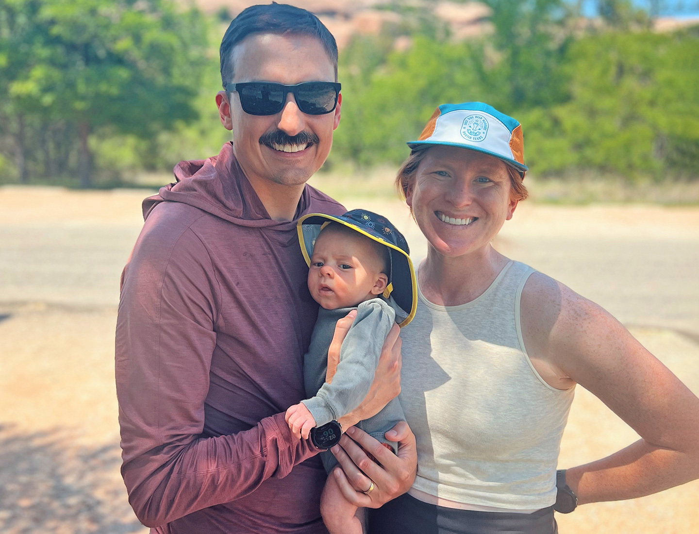 Katharina, her husband in sunglasses, and baby Elliott smile in a sunny outdoor family photo.