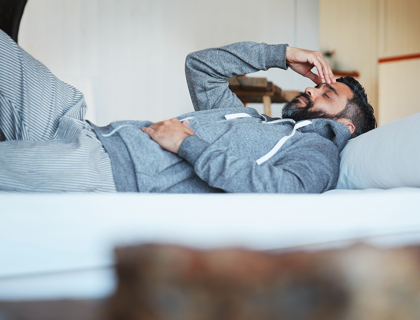 A man with short black hair and a beard is wearing gray sweatpants and a gray hoodie is laying on his bed with his hand on his forhead with his eyes closed. He looks fatigued. image