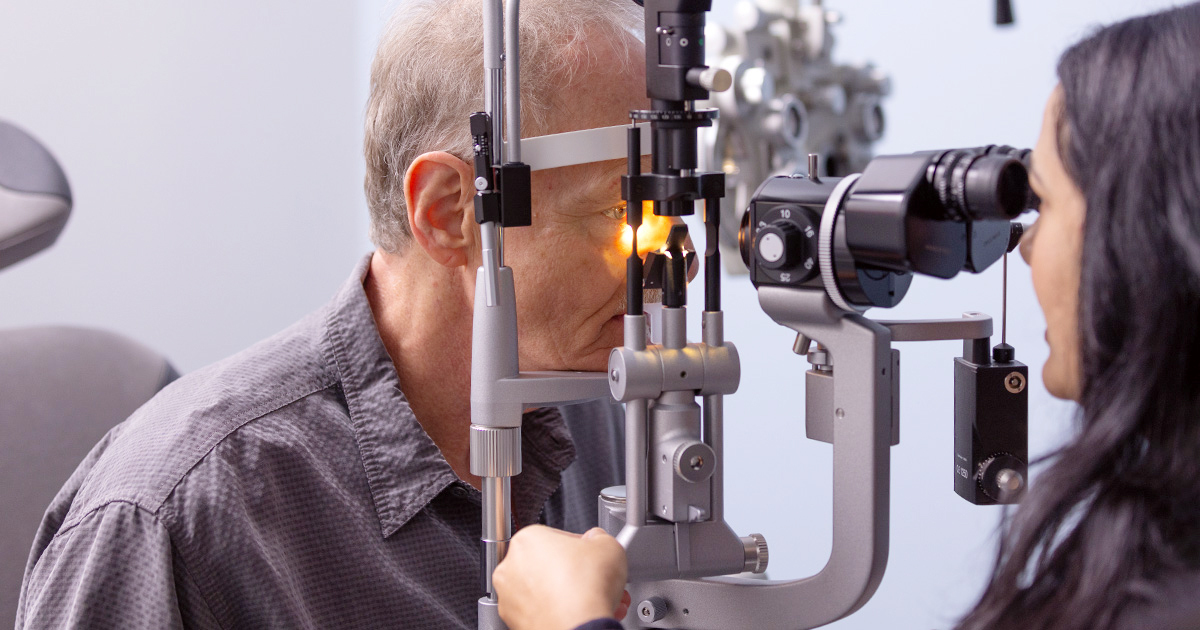 An older adult man receives an eye exam as a provider looks through a slit lamp microscope to assess vision.
