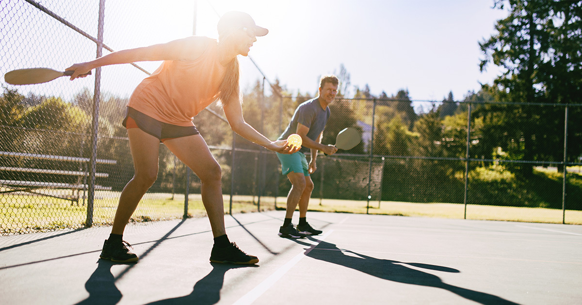 Two people playing pickleball on an outdoor court, with one player lunging forward to hit the ball, promoting tips for preventing common pickleball injuries.