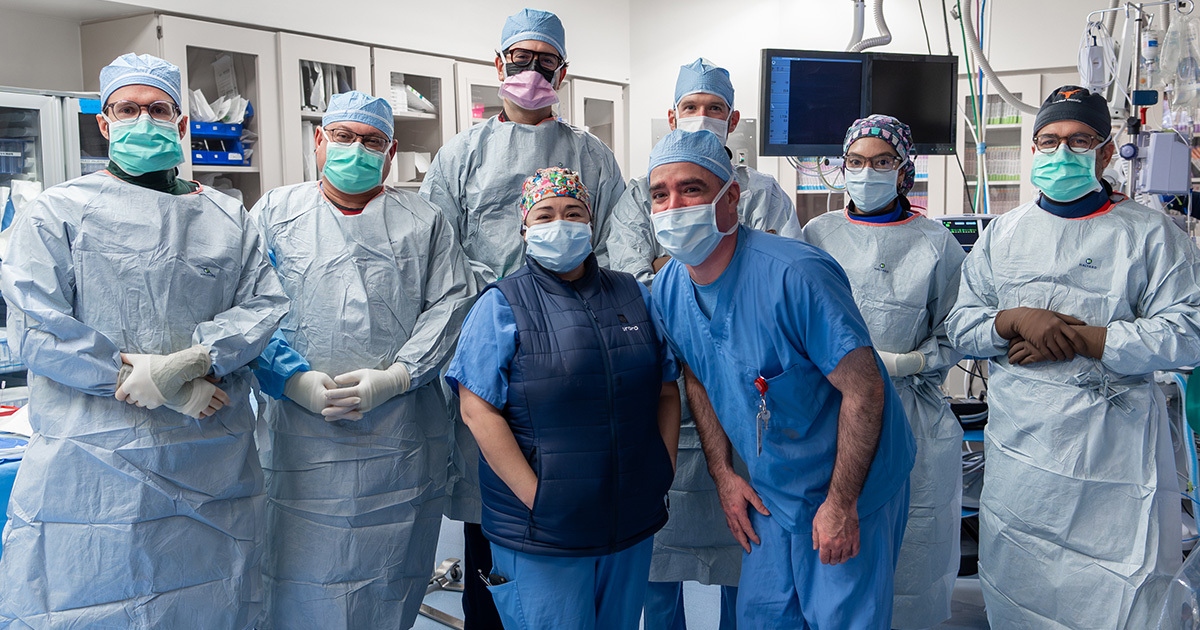 Cardiovascular care team in scrubs and masks pose together in a procedure room.