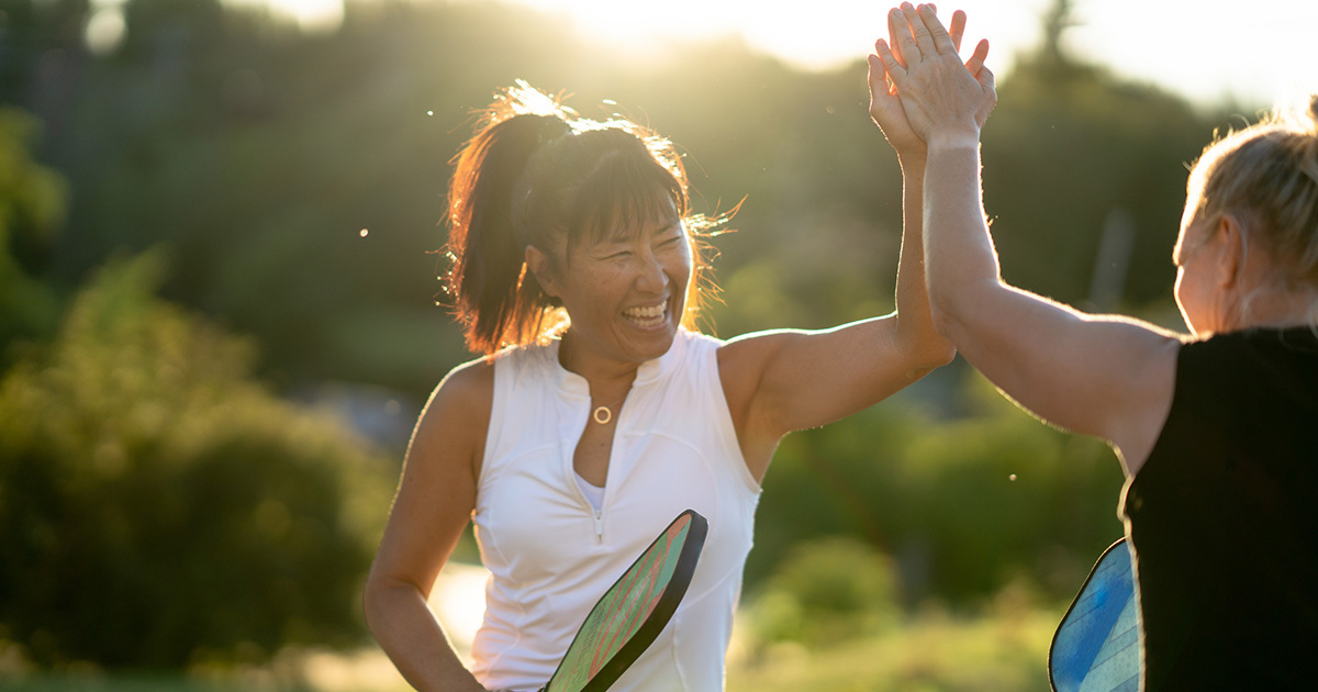 Two adult women in athletic wear high-five while holding pickleball paddles outdoors in the late afternoon sun.