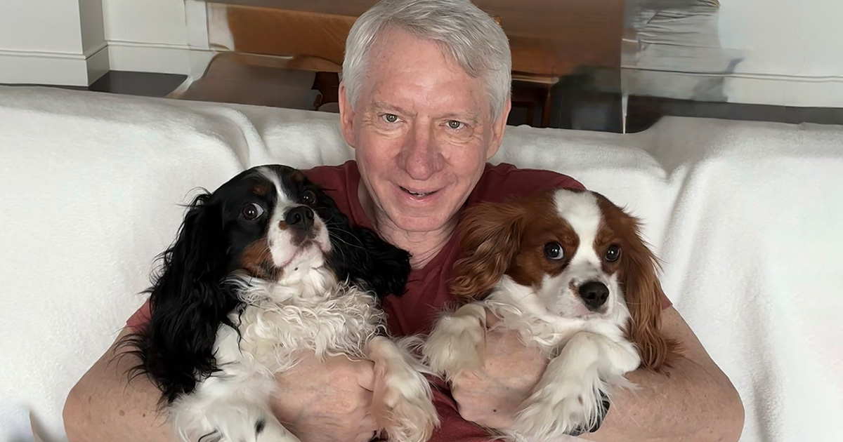 Smiling Donald “Don” Kettl, with short white hair sitting on a couch, holding two Cavalier King Charles Spaniels — one with black and white fur and one with chestnut and white fur.