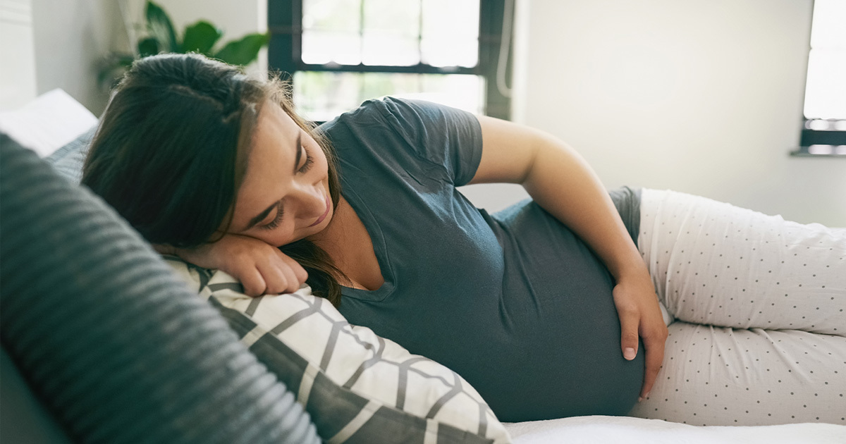 A pregnant woman lying on her right side on a couch looking down and holding her belly with her left hand.