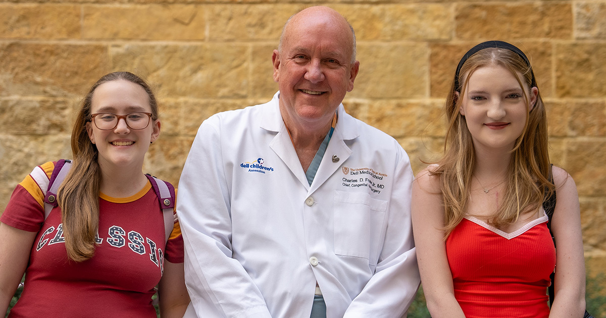Two smiling young women stand on either side of a doctor in a white coat, all posing happily in front of a stone wall.