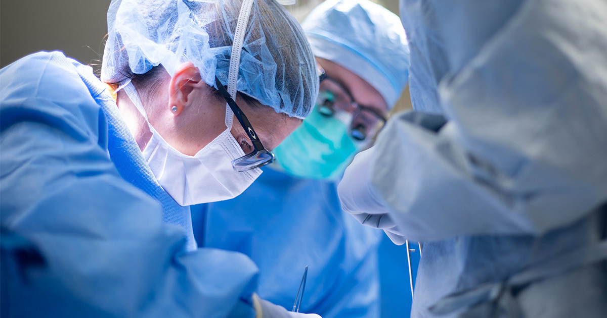 Dr. Nicole Turgeon performing a procedure in the operating room as another team member looks on. Both are wearing surgical masks, head coverings, gloves, and gowns.