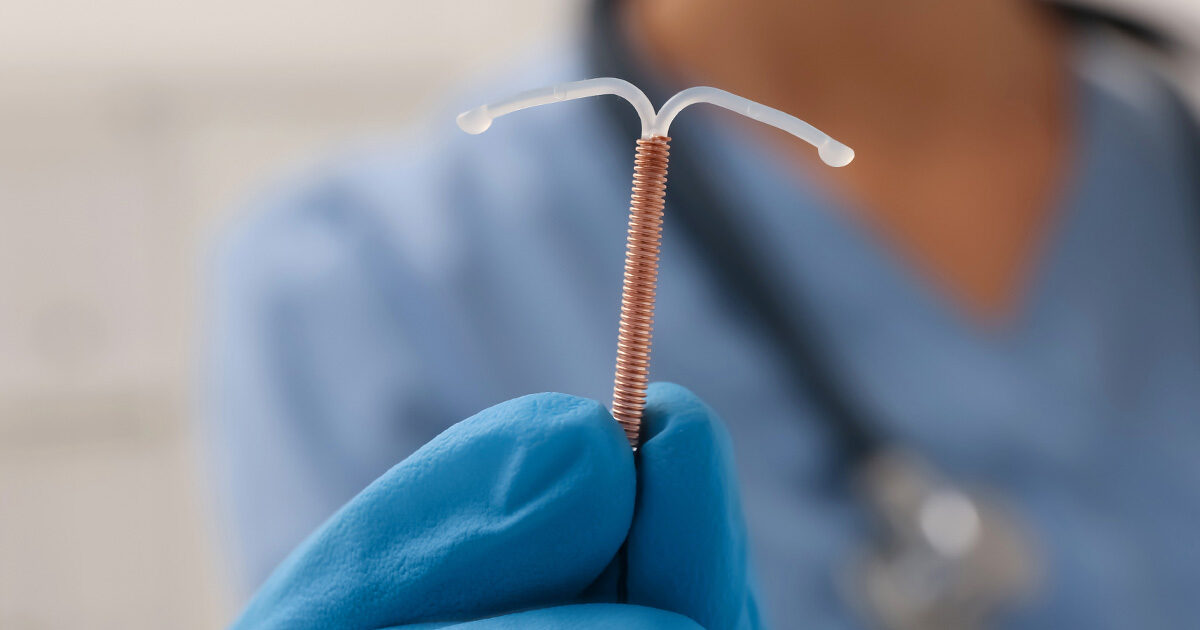 An IUD device is close up and in focus while a person wearing a stethoscope, surgical gloves, and blue scrubs is blurry in the background.