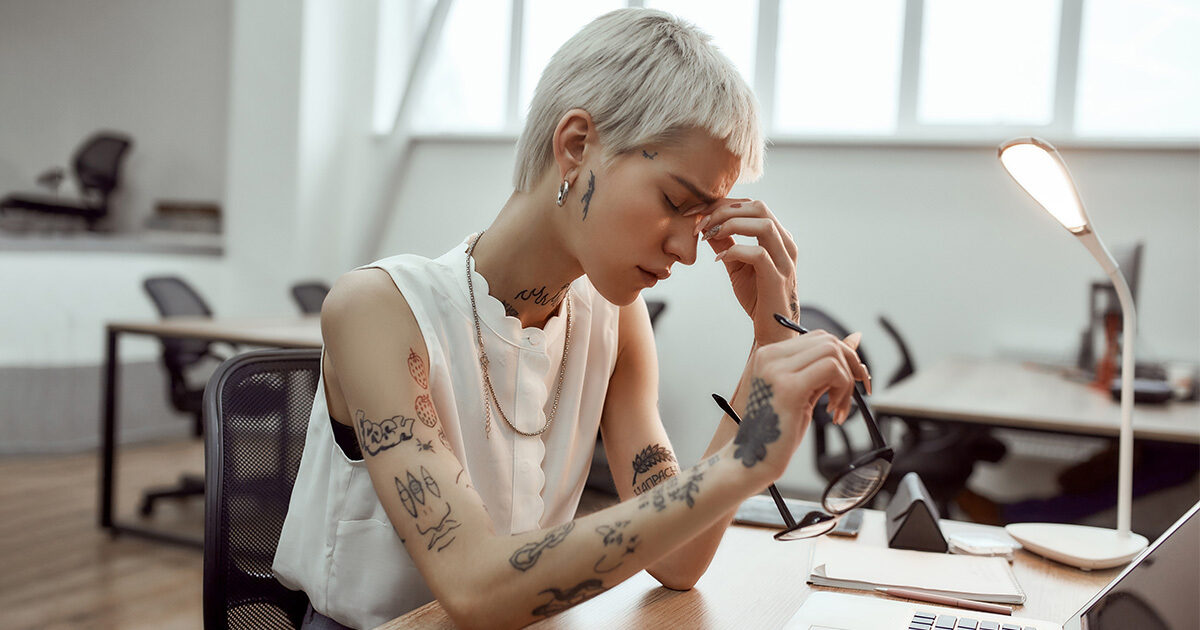 A woman sitting at a desk with a laptop squints and pinches the bridge of her nose, holding her glasses with her other hand.
