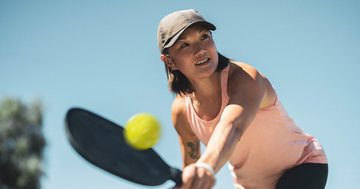 Adult woman in athletic wear and cap swings a paddle to hit a pickleball outdoors on a sunny day.