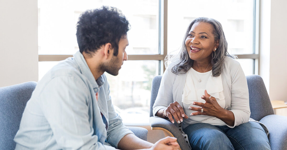 A young man and a woman with gray hair seated in blue chairs engaging in conversation.