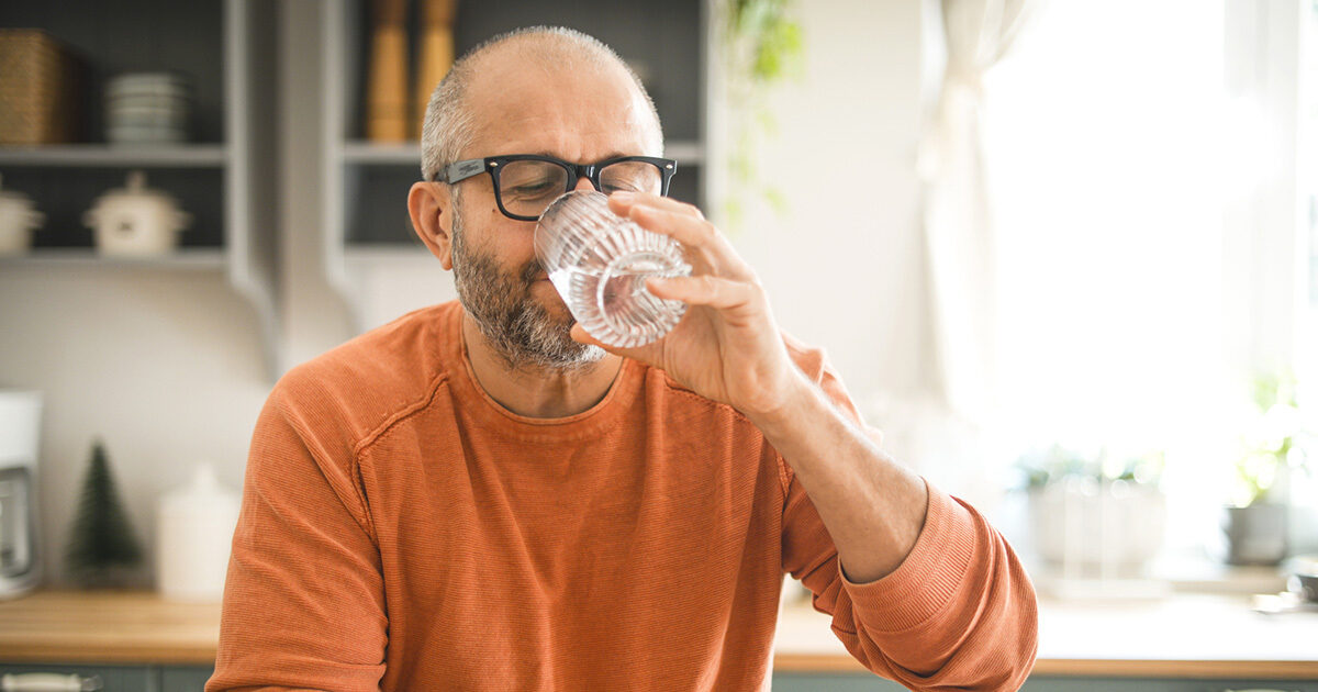 A man in an orange shirt drinks a glass of water in his kitchen.