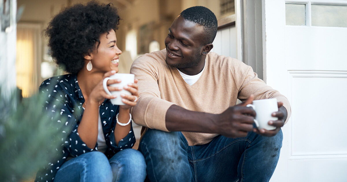 Two adults sit on a porch smiling at each other while holding coffee mugs in a relaxed conversation.