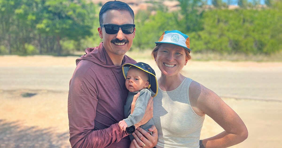 Katharina, her husband in sunglasses, and baby Elliott smile in a sunny outdoor family photo.