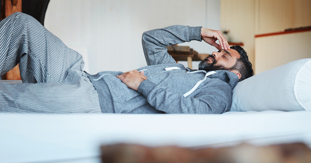 A man with short black hair and a beard is wearing gray sweatpants and a gray hoodie is laying on his bed with his hand on his forhead with his eyes closed. He looks fatigued.