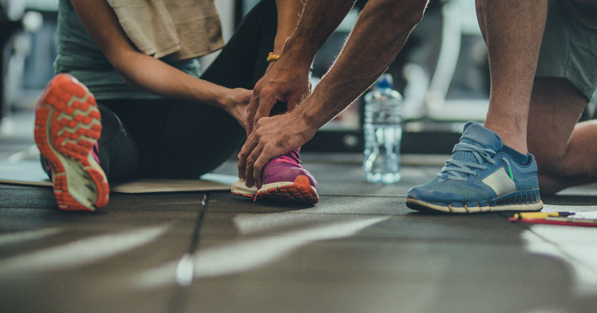 A closeup view of two people's feet, wearing athletic shoes. in a gym. One person is sitting on the floor while the other leans over and places their hands on the person's foot and ankle.