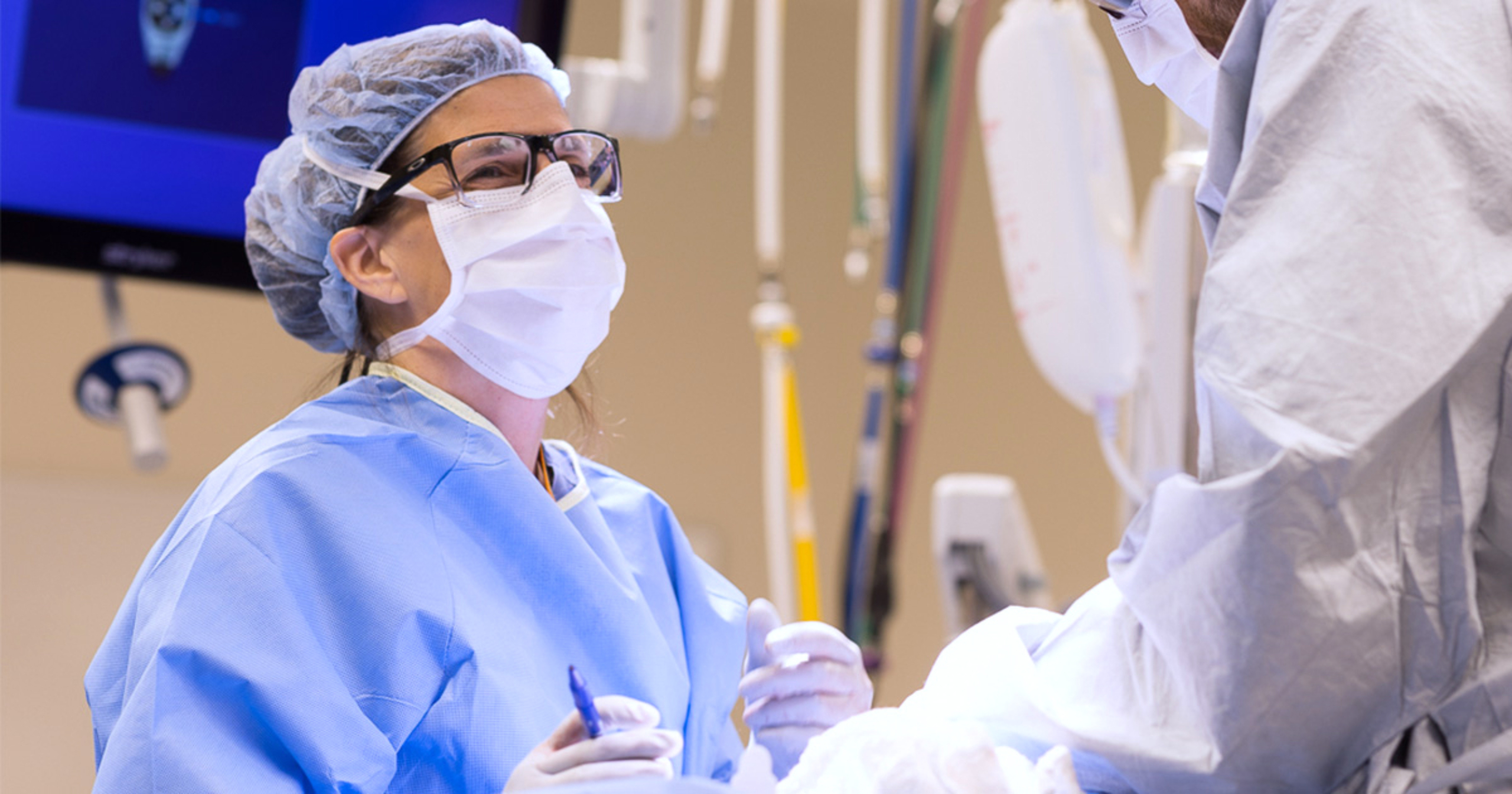 Dr. Nicole Turgeon speaking to another practitioner in the operating room. She is wearing a surgical mask, head covering, gown, and gloves, and is holding a surgical instrument in her right hand.