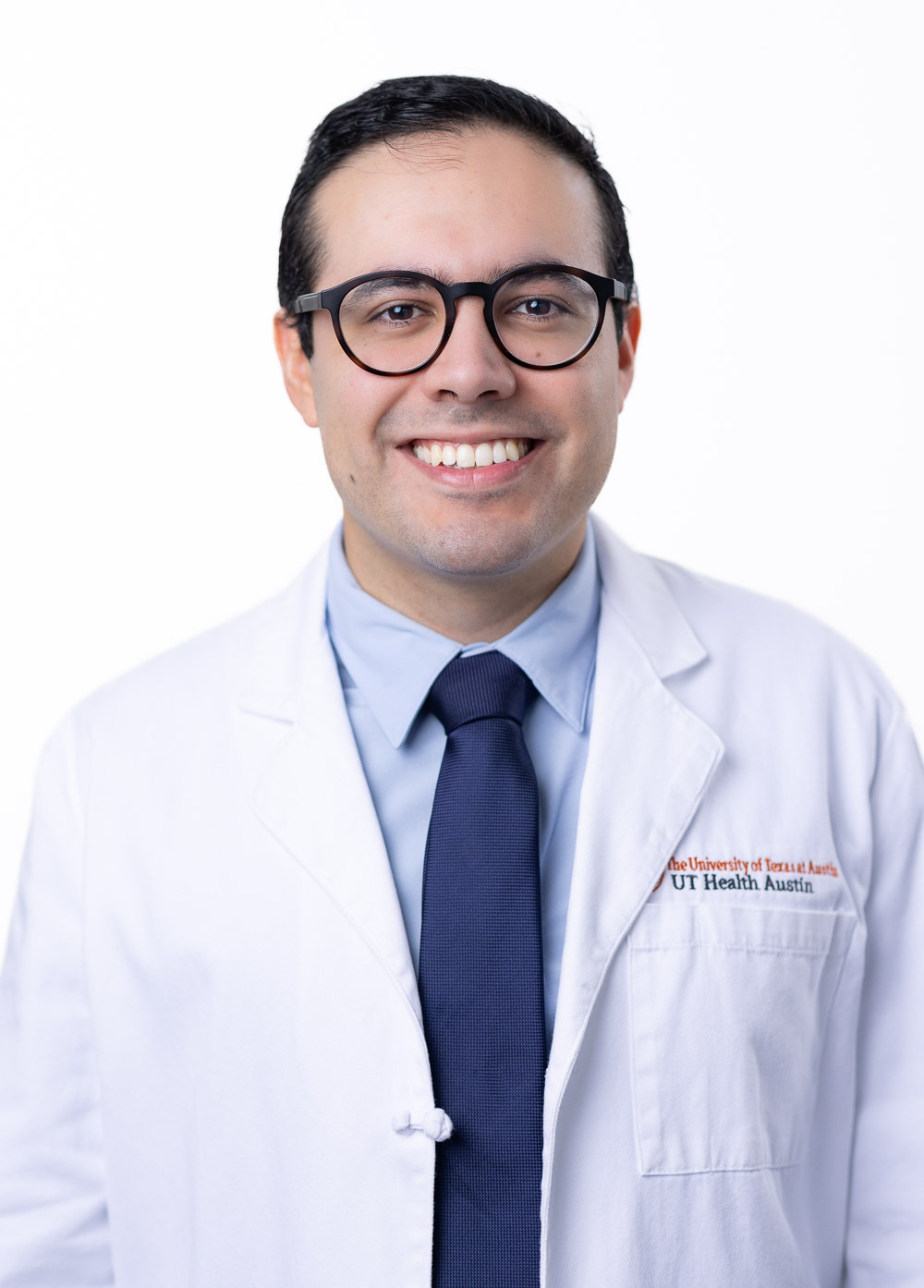 Felipe Flores is smiling at the camera.  He is wearing a white coat, light blue shirt, and a navy blue tie.