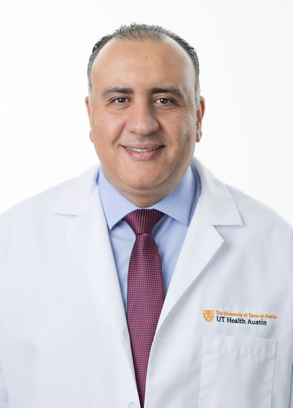 Dr. Mohammad Alsumrain is smiling at the camera and is wearing a white coat, blue shirt, and maroon tie.