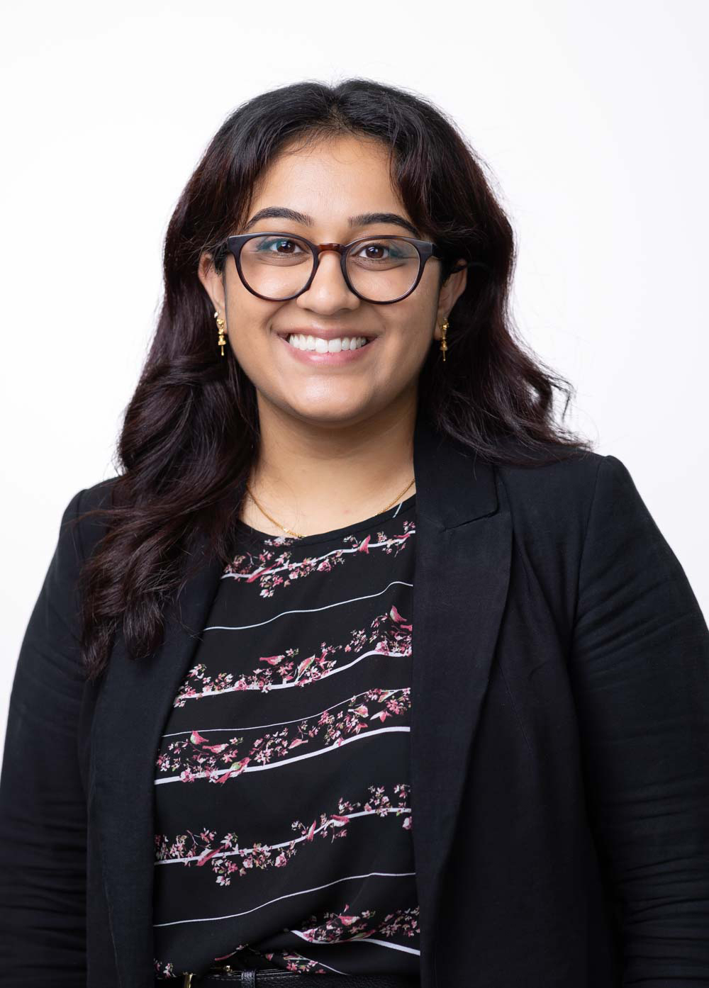 Ramya Srikanth is smiling and looking at the camera. She has black glasses and is wearing a black and white striped blouse with pink flowers on it, along with a black blazer.
