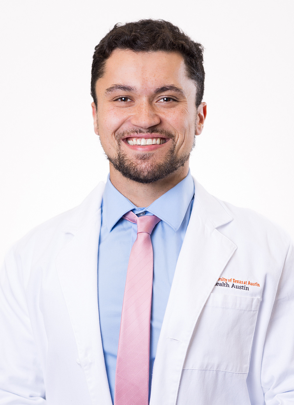 Thomas Ratana is smiling at the camera and wearing a white coat, light blue shirt, and a pink tie.
